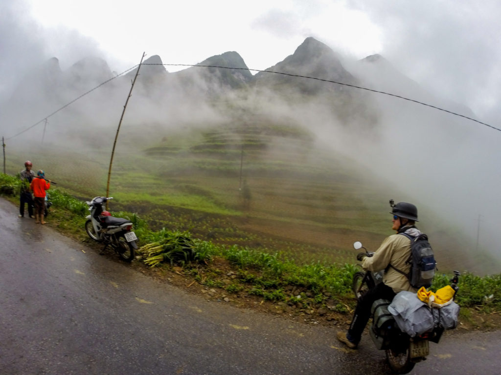 Motorbikes parked beside a rice field in Vietnam, with majestic karst mountains shrouded in heavy fog in the background.