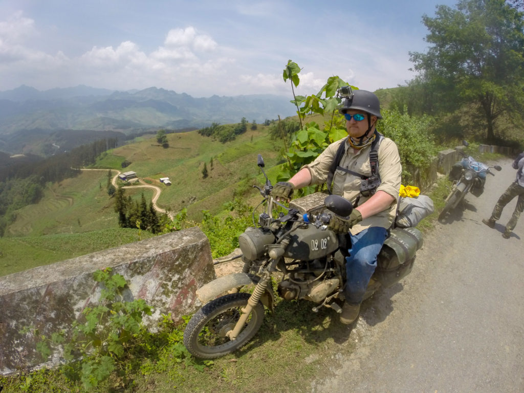 Rider on a customized Minsk motorcycle, pausing on a rugged trail amidst Vietnam's breathtaking mountains and valleys.