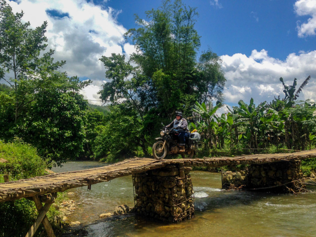 Motorcyclist crossing a rustic bridge surrounded by lush greenery on one of the best Vietnam motorbike tours with Motorbike Adventures Vietnam.